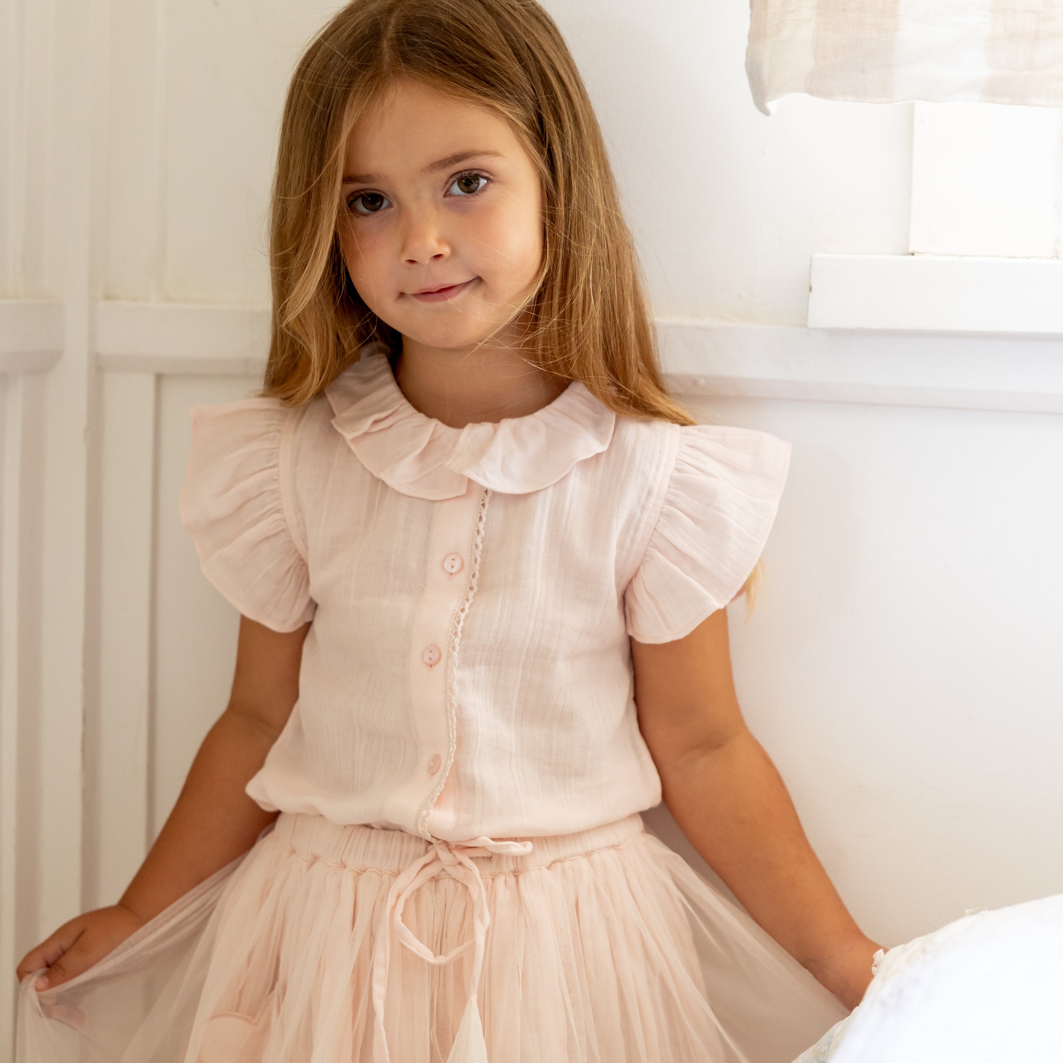 Young girl in a light pink dress sitting on a bed in a softly lit room.