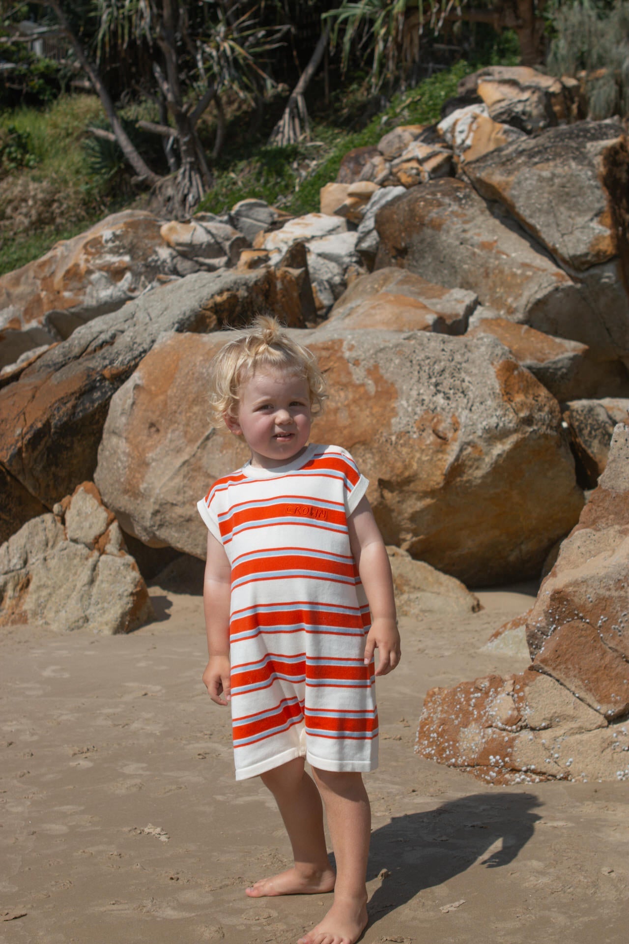 Child in a red and white striped romperstanding on a rocky beach.