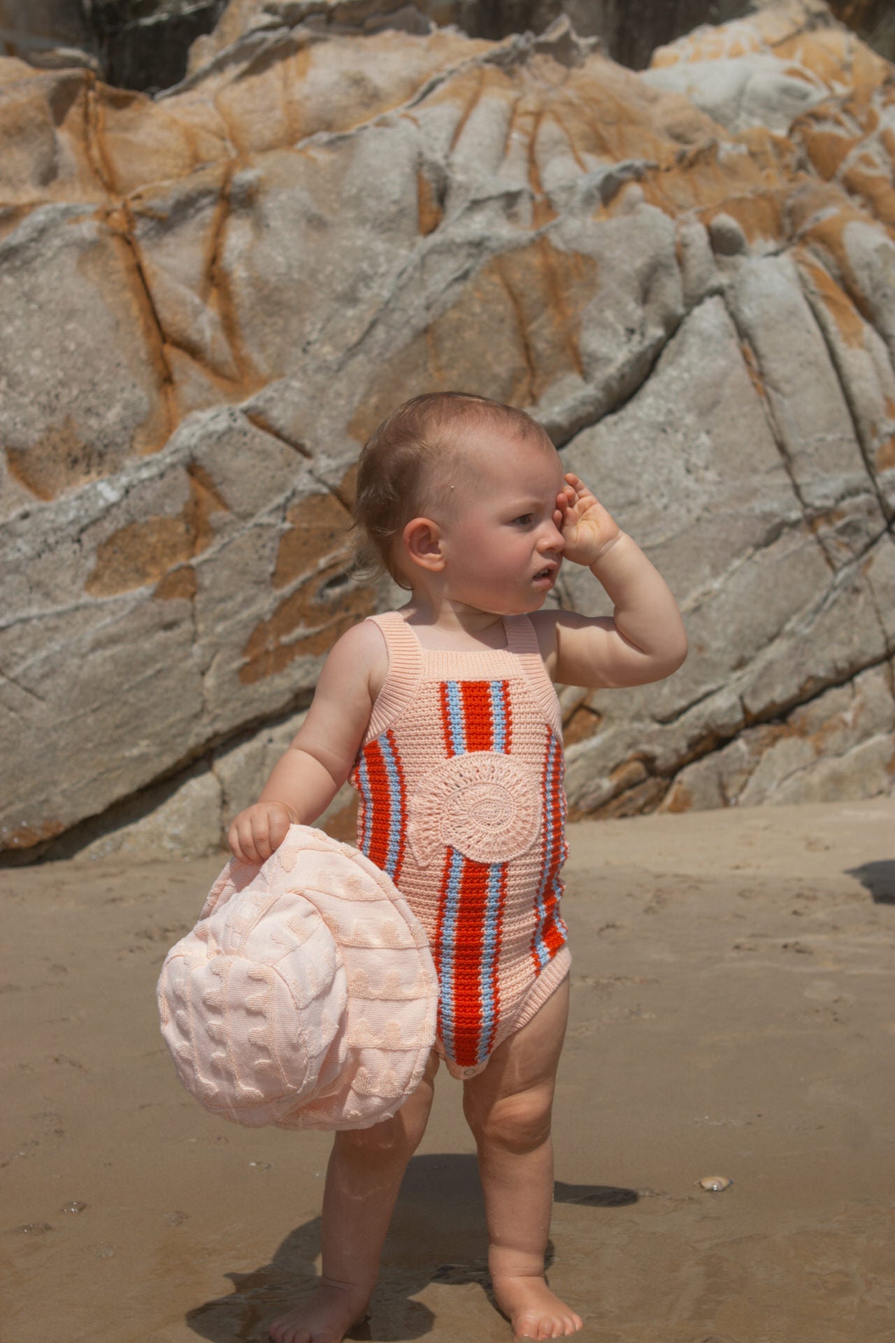Baby in a striped singletsuit standing on a beach with rocks in the background