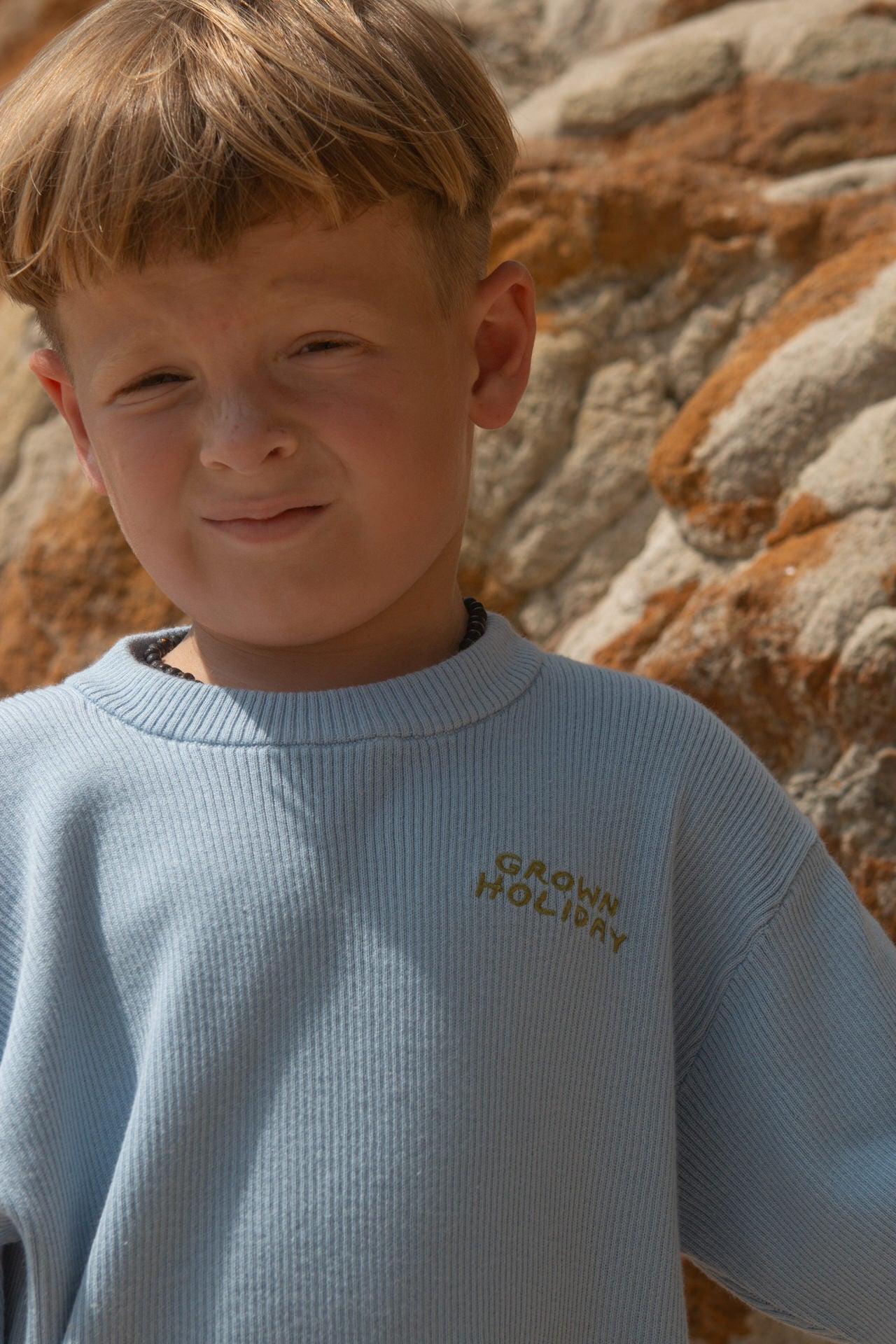 Young boy wearing a light blue tee with text, standing against a rocky background